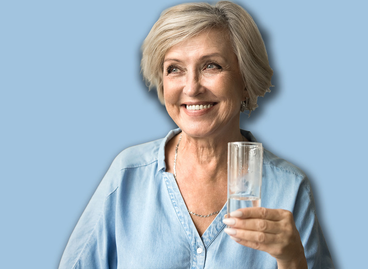 Happy satisfied elderly lady holding transparent glass pf pure clean water, standing in home interior, looking away with toothy smile, enjoying good hydration balance