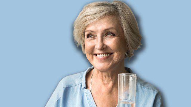 Happy satisfied elderly lady holding transparent glass pf pure clean water, standing in home interior, looking away with toothy smile, enjoying good hydration balance