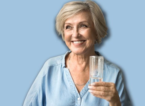 Happy satisfied elderly lady holding transparent glass pf pure clean water, standing in home interior, looking away with toothy smile, enjoying good hydration balance