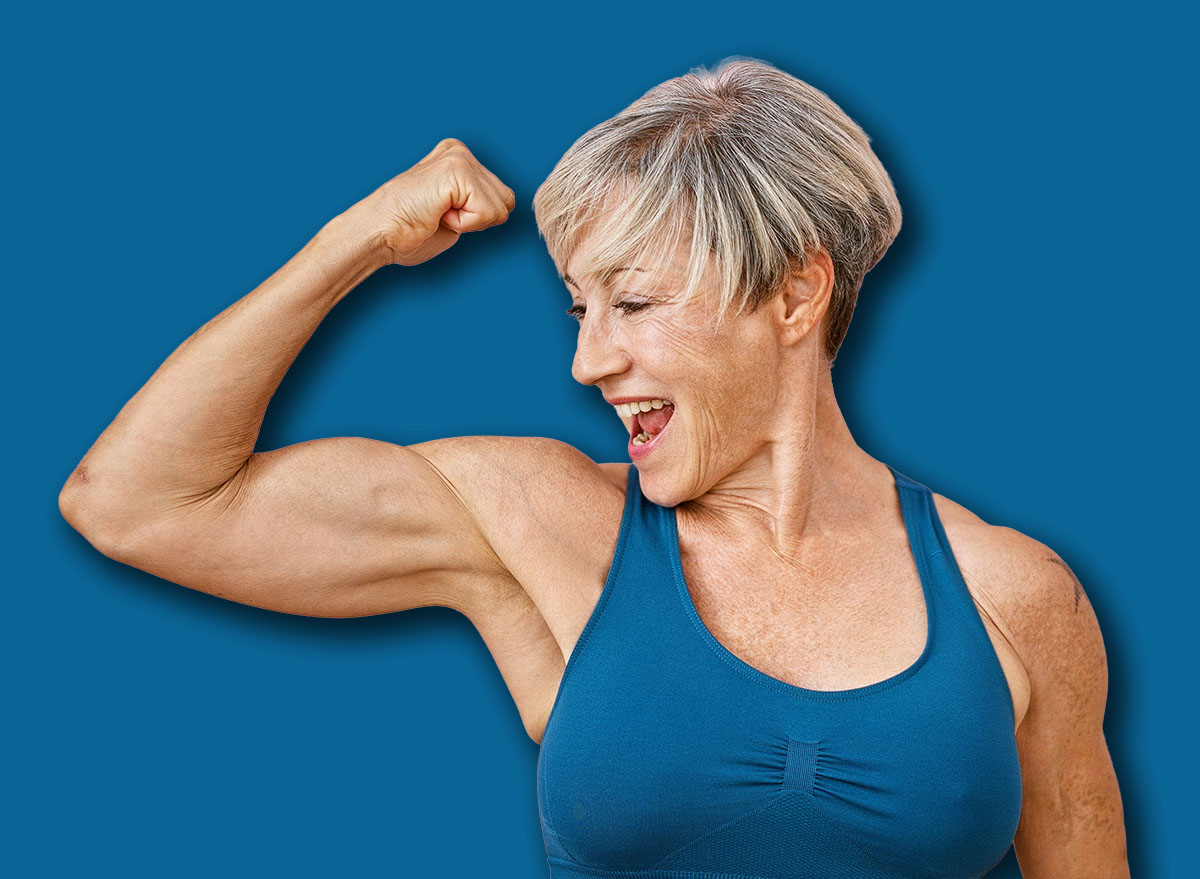 A fit, mature woman with short grey hair flexes her muscles against a pink isolated background, displaying strength and vitality.