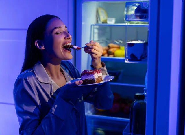 Happy woman eating sweet cake late at night, standing near opened fridge and holding plate with dessert, feeling satisfied
