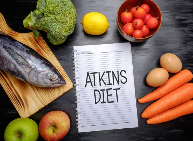 Atkins Diet on chalkboard, health conceptual. Healthy fresh food fish, lemon, tomatoes, apple, carrot and broccoli.