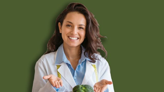 Portrait of cheerful dietitian in lab coat pointing at fresh fruits and vegetables on table, smiling at camera at clinic. Weight loss consultant recommending healthy plant based diet