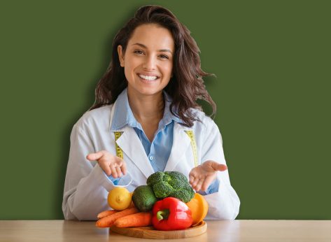 Portrait of cheerful dietitian in lab coat pointing at fresh fruits and vegetables on table, smiling at camera at clinic. Weight loss consultant recommending healthy plant based diet