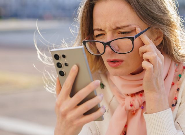 Woman struggling to read phone screen outdoors, squinting through glasses, showing symptoms of poor eyesight and digital fatigue.