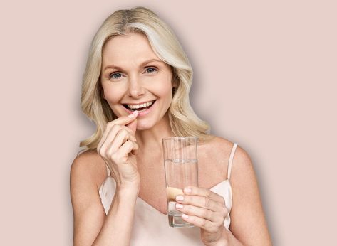 Happy middle aged 50s woman holding pill and glass of water taking dietary supplements. Portrait of smiling adult attractive woman taking care of health in menopause, isolated on white.