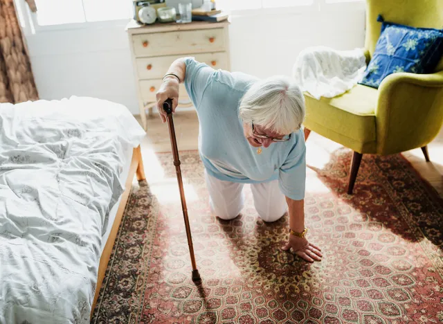 Elderly woman with white hair using a cane, kneeling on a patterned rug in a bright room. The elderly woman appears to be in a cozy, well-lit space. Old woman falling to the floor in care home.