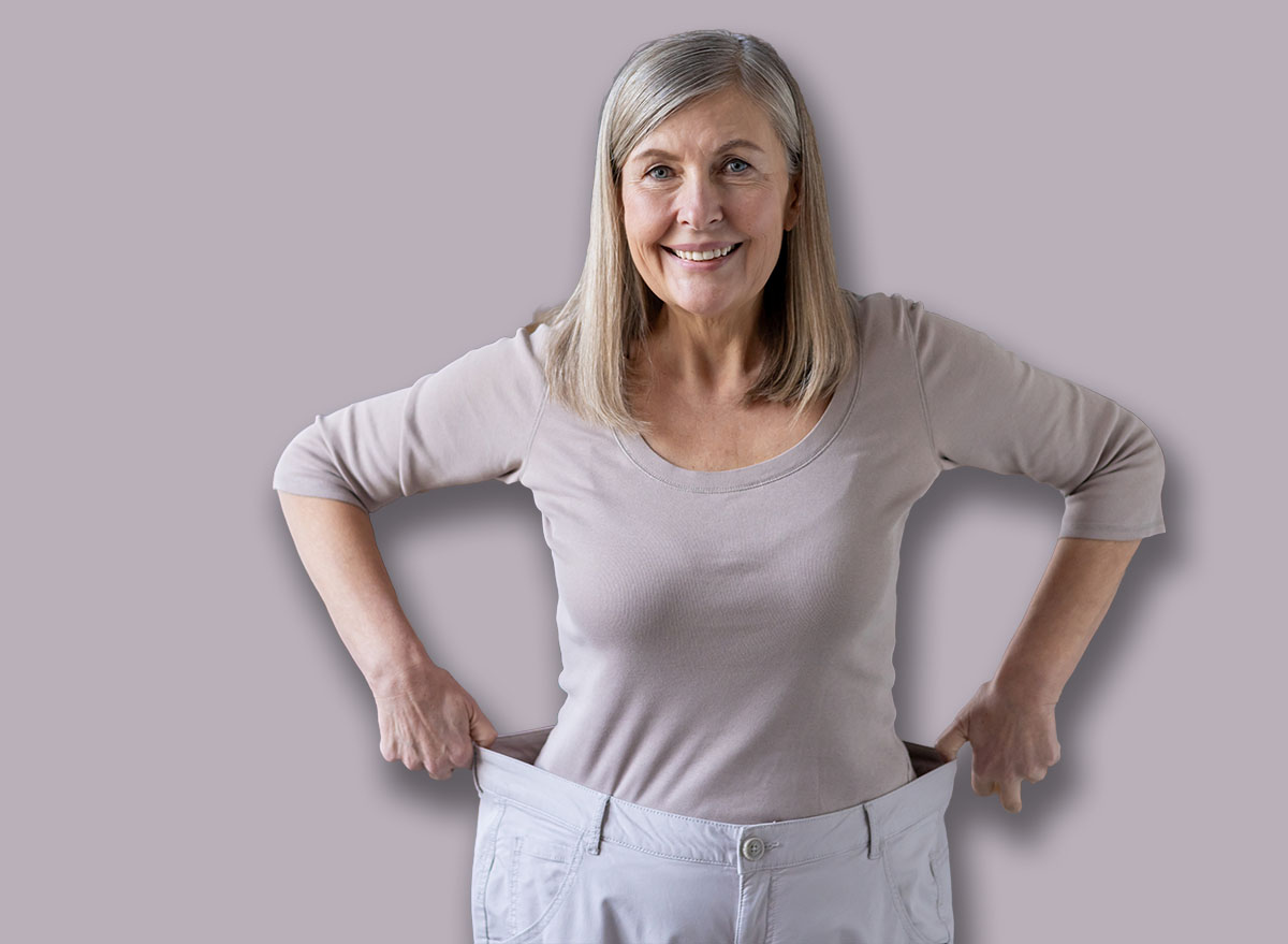 Joyful mature woman stands confidently showing weight loss wearing oversized pants, expressing happiness and success. Her smiling face reflects pride in personal health and fitness accomplishment.
