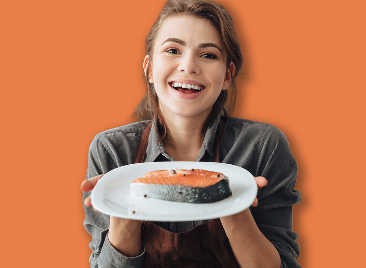 Image of young happy woman standing in kitchen while cooking fish. Looking at camera.