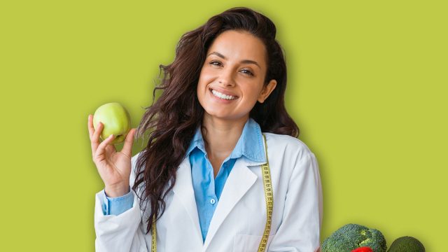 Cheerful female nutrition adviser with green apple and tape measure smiling at camera, standing at clinic office, free space. Weight loss specialist promoting healthy eating habits