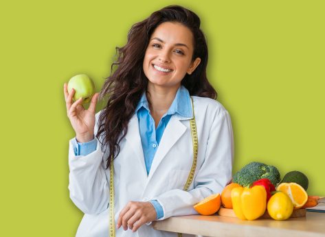 Cheerful female nutrition adviser with green apple and tape measure smiling at camera, standing at clinic office, free space. Weight loss specialist promoting healthy eating habits