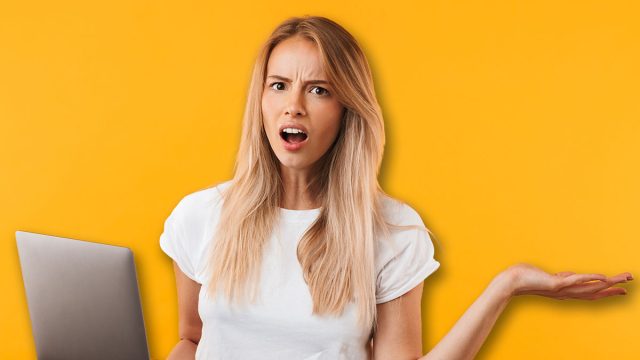 Portrait of a confused young blonde girl holding laptop computer isolated over yellow background