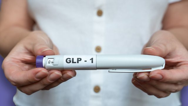 Close-up of a woman holding a GLP-1 injection pen in both hands with her bare abdomen visible in the background. The image illustrates weight management, hormone therapy, modern medical treatment, and